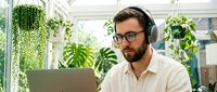 Man with dark hair and beard, wearing glasses and white shirt, working on laptop with headphones in bright office surrounded by green plants and natural light.