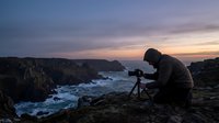 Male photographer capturing dramatic coastal sunset with professional camera on tripod overlooking rocky cliffs and ocean waves during golden hour twilight.