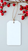 Blank white gift tag with twine string hanging near red winter berries on branches against light gray background, mockup for holiday greeting or price label.