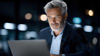 Confident mature businessman with gray beard working on laptop in modern office with city lights background for corporate presentations.