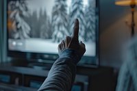 Person pointing finger at TV screen showing winter forest scene with snow-covered trees, creating interactive viewing experience in dark room atmosphere.