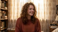 Young woman with curly brown hair smiling warmly in cozy home library wearing rust colored sweater. Natural lighting creates inviting atmosphere for lifestyle content.