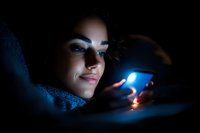 Young woman with curly hair illuminated by phone screen light in dark room, creating dramatic blue glow on face while browsing social media at night.