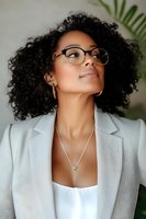 Young African American businesswoman with natural curly hair wearing glasses and white blazer looking up confidently against tropical plant background.