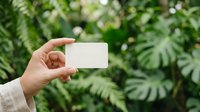 Person with medium skin tone holding blank white card against lush green foliage background, suitable for business branding, eco-friendly marketing, or nature-related promotional materials.