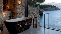 Luxurious black marble bathtub in modern cave bathroom with panoramic sea view, coastal cliffs, and ambient lighting for relaxing spa experience.