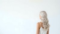 Mature woman with long silver wavy hair wearing white sleeveless top, standing in profile against neutral background, suitable for beauty, wellness, and lifestyle design projects.