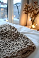 Cozy knitted blanket on white bedding with dried flowers in vase and winter window view, creating warm atmospheric evening mood with soft bokeh lights.