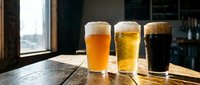 Three different beer glasses with wheat, lager and stout varieties on wooden table in modern pub interior with natural window lighting for beverage marketing.