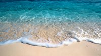 Crystal clear turquoise ocean waves gently rolling onto sandy beach with white foam, creating natural pattern where water meets pristine shoreline in tropical paradise.