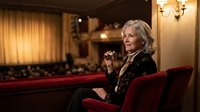 Elegant senior Caucasian woman with silver hair sitting in luxurious theater seat, wearing black formal attire, sophisticated cultural entertainment venue.