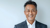 Confident mature Asian businessman in dark suit and white shirt smiling warmly against light background, expressing success and leadership in corporate setting.