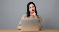 Young Hispanic businesswoman in beige sweater thinking while working on laptop at wooden desk, contemplating business decisions in modern office setting.