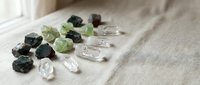 Collection of raw gemstones and crystals including green peridot, dark tourmaline, and clear quartz specimens arranged on neutral linen fabric surface.