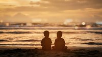 Two young children sitting together on beach at golden sunset watching ocean waves with mountains in background creating peaceful family moment.