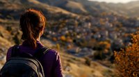 Young woman with backpack enjoying scenic mountain sunset view over small town in valley, warm autumn colors illuminate peaceful landscape at golden hour.