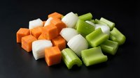 Fresh diced vegetables including orange carrots, white onions and green celery stalks chopped into small cubes on dark surface for cooking preparation.