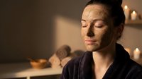 Young Caucasian woman enjoying relaxing spa treatment with natural clay face mask in candlelit wellness center for skincare and beauty routine.