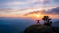 Person celebrating success with arms raised on mountain peak during golden hour sunrise with dramatic cloudy sky and layered mountain ranges in background.