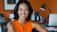 Asian woman in bright orange sleeveless top smiling confidently at camera in modern office setting with desk lamp and workspace background. Professional corporate portrait.