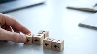 Wooden blocks with arrows pointing in different directions arranged on a desk, with a hand placing one block, symbolizing choice and strategy.