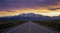 Straight asphalt highway stretching toward snow-capped mountain peak at sunset, dramatic purple and golden sky, desert landscape with sparse vegetation, travel and adventure concept.