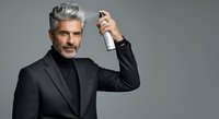 Mature man with gray hair and facial hair, wearing black business suit and turtleneck, holding hair spray bottle against neutral background for grooming and styling concepts.