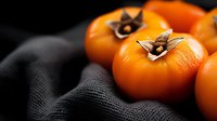 Fresh ripe persimmons with brown calyxes on dark textile background, selective focus. Seasonal autumn fruits in moody lighting for food photography.