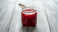 Homemade red jam or jelly in glass jar with spoon on wooden table surface, ready for breakfast or baking use.