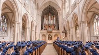 Graduation ceremony in majestic Gothic cathedral with students in blue caps and gowns seated in wooden pews beneath soaring arches and ornate pipe organ.
