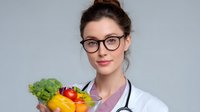 Young female doctor in white coat and glasses holding fresh vegetables and fruits in glass bowl promoting healthy eating and nutrition against gray background.