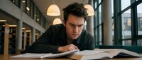 Young focused Caucasian male student studying intensively at library table with open books. Academic learning and education concept for university preparation.