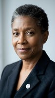 Professional African American businesswoman with short gray hair in dark suit jacket looking confidently at camera in corporate office setting.