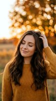 Young woman with long wavy brown hair wearing mustard yellow sweater touching her head with eyes closed in peaceful moment during golden hour sunset.