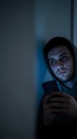 Man in dark hoodie holding smartphone in moody blue lighting, depicting cybercrime, hacking, online fraud, digital theft, and internet security threats for crime prevention campaigns.
