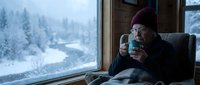 Elderly Caucasian woman in purple knit hat enjoying hot beverage by large window overlooking snowy winter forest landscape from cozy indoor setting.