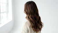 Young woman with long wavy brown hair wearing beige knit sweater standing by window in bright modern interior for lifestyle and fashion content.