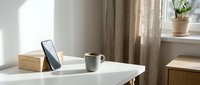 Minimalist workspace with smartphone, coffee mug, and wooden accessories on white desk near window with natural light and curtains.