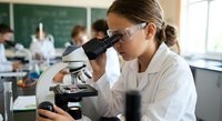 Young female student in safety goggles using microscope in science laboratory classroom for educational research and scientific study.