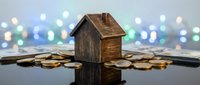 Miniature wooden house model sitting on scattered coins with colorful bokeh lights background representing real estate investment and savings concept.