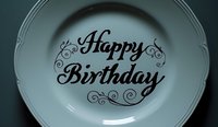 Elegant white ceramic plate with decorative Happy Birthday text in black calligraphic lettering and ornate swirl details against soft blurred background.