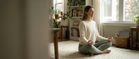 Young Caucasian woman practicing meditation in lotus pose on carpet in bright living room with plants and natural light for mindfulness and wellness.
