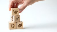 Hand stacking wooden blocks with gardening icons including watering can, flower, shovel, sun and leaf symbols for eco-friendly agriculture concepts.