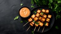 Grilled tofu skewers with spicy sauce on black plate garnished with fresh herbs. Asian fusion appetizer served with creamy dipping sauce on dark background.