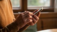 Person in cozy sweater using smartphone at home by window with warm natural lighting for communication and digital lifestyle.