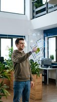 Young Caucasian man interacting with holographic digital interface in modern eco-friendly office with plants and natural lighting for technology presentations.