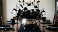 Elegant dining room table set with dark floral centerpiece in metallic bowl, black candles, and formal place settings creating sophisticated atmosphere.