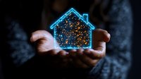 Glowing blue house silhouette with sparkles floating in cupped hands against dark background, representing real estate dreams, home ownership and property investment.