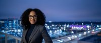 Professional woman with curly dark hair and glasses wearing black blazer standing on rooftop overlooking illuminated cityscape at dusk for corporate and business concepts.