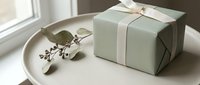 Elegant sage green gift box tied with a cream ribbon beside a eucalyptus sprig on a neutral tray, soft natural light, minimalist style.